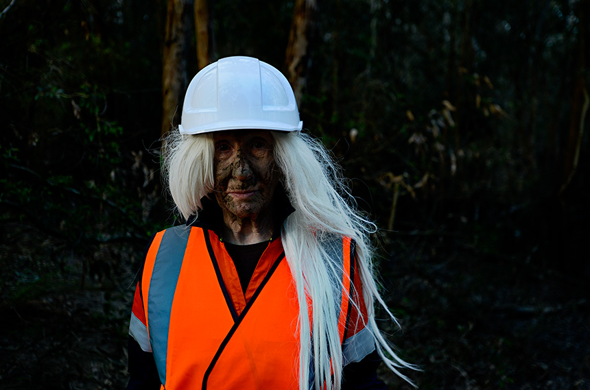Jill Orr dressed as a dirty faced worker, in high vis and a hard hat standing in a river.